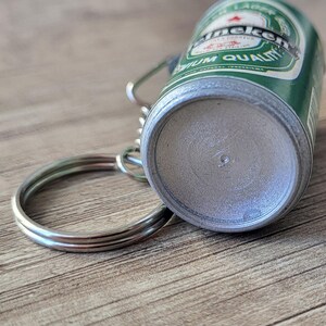 May include: A miniature Heineken beer can keychain. The green and silver can features the Heineken logo and text. A silver key ring is attached to the top of the can.