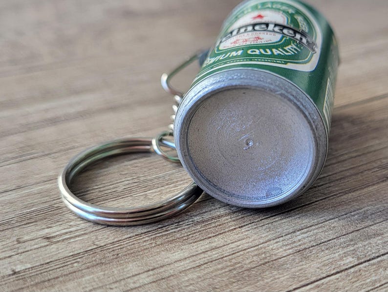 May include: A miniature Heineken beer can keychain. The green and silver can features the Heineken logo and text. A silver key ring is attached to the top of the can.