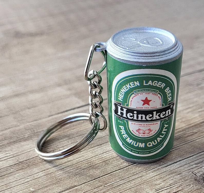 May include: A green Heineken Lager Beer can keychain with silver accents. The keychain features the Heineken logo and text on the can. The keychain is attached to a silver chain and ring.