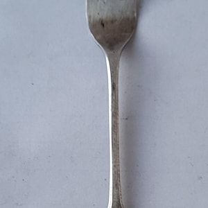 Puede incluir: Un tenedor de metal plateado con tres dientes largos y delgados y un mango plano y cónico. El mango muestra dos pequeños sellos rectangulares estampados cerca del cuello, que indican la calidad o el origen del metal. Acabado mate.