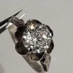 Puede incluir: Un anillo de tono plateado con una gran piedra redonda transparente facetada engastada en el centro de una montura metálica en forma de flor. El aro del anillo está decorado con patrones de hojas en relieve a ambos lados de la montura floral.