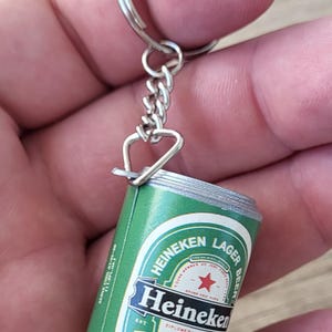 May include: Miniature Heineken Lager Beer can keychain. The green and white can features the Heineken logo and text. The keychain has a silver-colored ring and chain.
