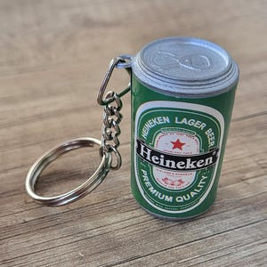 May include: A miniature Heineken Lager Beer can keychain. The green and white can features the Heineken logo and text. The keychain has a silver key ring and chain.