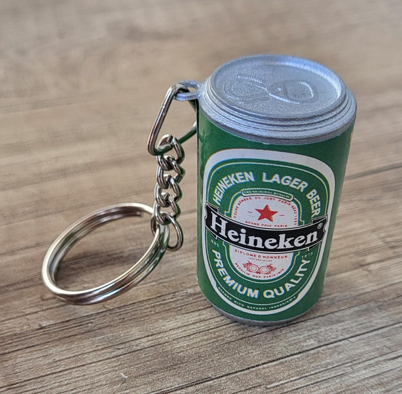 May include: A miniature Heineken Lager Beer can keychain. The green and white can features the Heineken logo and text. The keychain has a silver key ring and chain.