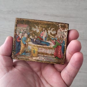 Puede incluir: Pequeño icono religioso rectangular y antiguo con una ilustración detallada de una escena bíblica. La obra de arte representa figuras con túnicas coloridas alrededor de una figura central en una cama, con ángeles y elementos arquitectónicos en el fondo.