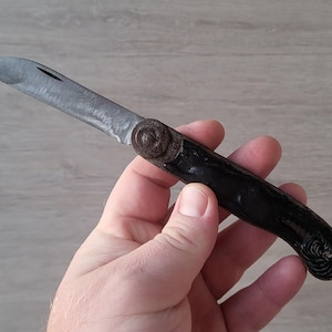 May include: A folding pocket knife with a single silver-toned metal blade and a dark, textured handle. The pivot point features a circular, weathered metal detail, and the handle has a curved shape with intricate, dark embossed patterns at the end.