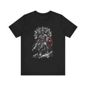 Puede incluir: Camiseta negra con una ilustración detallada de un guerrero vikingo. El gráfico es en tonos de blanco, gris y rojo, representando al guerrero con un escudo y una espada. El diseño incluye runas nórdicas y un emblema circular.