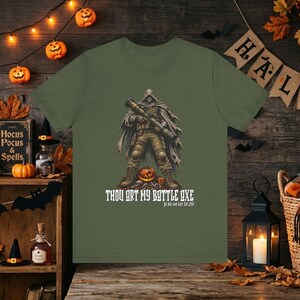 May include: An olive green t-shirt featuring a graphic of a hooded figure in tactical gear holding a rocket launcher, standing over a jack-o'-lantern. The text below reads, "THOU ART MY BATTLE AXE JEREMIAH 51:20" in a white, stylized, gothic-inspired font.