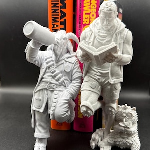 Könnte beinhalten: Ein Set aus drei unbemalten 3D-gedruckten Figuren, darunter eine ziegenartige Figur, die aus einer Flasche trinkt, eine menschliche Figur, die ein offenes Buch liest, und ein kleines Wesen mit Schutzbrille, vor Büchern mit den Titeln Matt Dinniman und Dungeon Crawler.