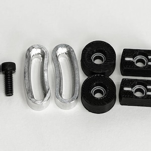 May include: An assortment of hardware components, including two black bolts, two silver-colored curved pieces, two black circular pieces, and two black rectangular pieces. The items are arranged on a white surface.