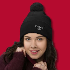 May include: Black beanie with a pom-pom on top. The beanie has the words "Dialysis Squad" embroidered in white. The background is a solid red color.