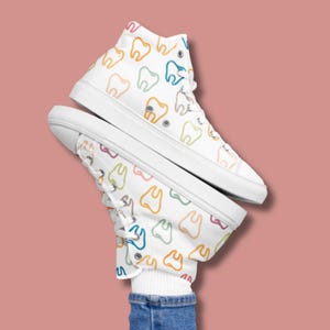 May include: White high-top sneakers with a colorful tooth pattern. The shoes feature a white rubber sole and laces. The design includes teeth in various colors, including orange, blue, and green. The sneakers are shown against a pink background.