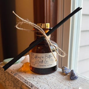May include: A brown glass diffuser bottle with a gold cap, black reeds, and a label that reads "Cute as a Button Candle Co. Coastal Cotton." The bottle is tied with twine and sits on a decorative surface with crystals.