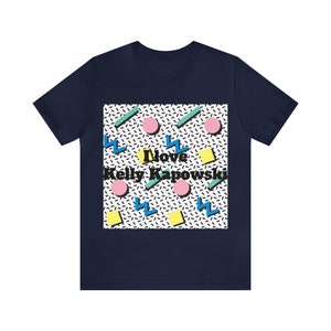 May include: A navy blue t-shirt with a white square featuring a geometric pattern in black, pink, yellow, blue, and green. The text "I love Kelly Kapowski" is printed in black.