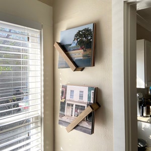 May include: Two vinyl record holders mounted on a wall. The holders are made of wood and are angled to display the records. The records are displayed vertically. The top record has a photo of a car and a person in a field. The bottom record has a photo of a building with a sign that says "Mumford and Sons".