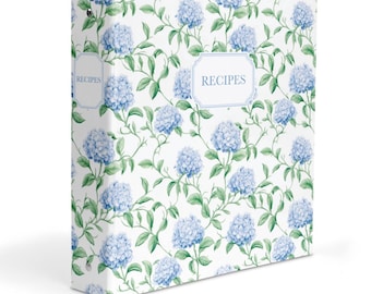 May include: A three-ring binder featuring a white background with a repeating pattern of blue hydrangea flowers and green leaves. The front cover and spine both display a rectangular label with a light blue border containing the word RECIPES.