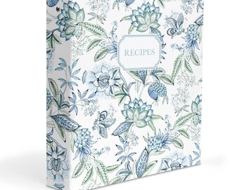 May include: A white three-ring binder featuring a repeating botanical pattern of blue and green flowers, leaves, and butterflies. The front cover displays a central rectangular label with a decorative border containing the word RECIPES in text.