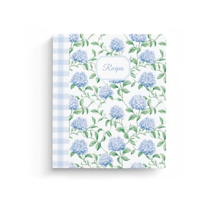 May include: A recipe book with a floral hydrangea design. The cover features blue hydrangeas and green leaves on a white background. The spine has a light blue gingham pattern. The word "Recipes" is in a white frame.