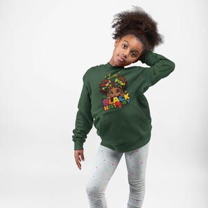 Könnte beinhalten: Ein junges Kind trägt ein grünes Hoodie mit dem Text "I am Black History" und einer Cartoon-Illustration eines jungen schwarzen Mädchens mit bunten Haarbändern. Das Hoodie hat eine Kängurutasche. Das Kind trägt graue Leggings mit einem weißen Herzmuster und rosa Turnschuhe.
