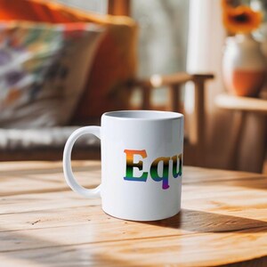 Può includere: Una tazza di ceramica bianca con un manico curvo. La parola "Equality" è stampata sul davanti a colori arcobaleno. La tazza è appoggiata su un tavolo di legno.