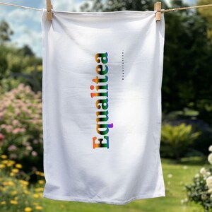 Può includere: Asciugamano bianco appeso a un filo con la parola "Equalitea" stampata verticalmente a colori arcobaleno. L'asciugamano è tenuto da mollette di legno su uno sfondo sfocato di fogliame verde e fiori.