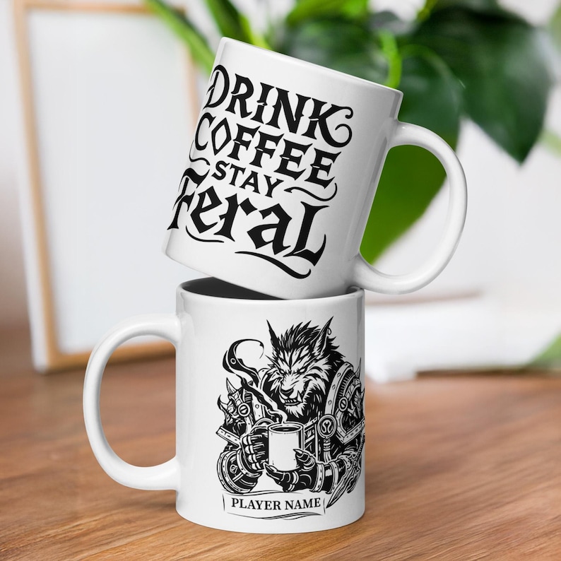 K&ouml;nnte beinhalten: Zwei wei&szlig;e Keramik-Kaffeetassen, die auf einer Holzoberfl&auml;che gestapelt sind. Auf der oberen Tasse steht in schwarzer Schrift "DRINK COFFEE STAY feral". Die untere Tasse zeigt eine schwarz-wei&szlig;e Illustration eines Werwolfs, der eine Tasse h&auml;lt, mit dem Text "PLAYER NAME" darunter.