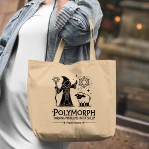 Può includere: Borsa tote beige con una grafica nera di un mago, una pecora e forme geometriche. Il testo sulla borsa recita "Polymorph Turning Problems Into Sheep" e "Player Name". La borsa ha due manici.