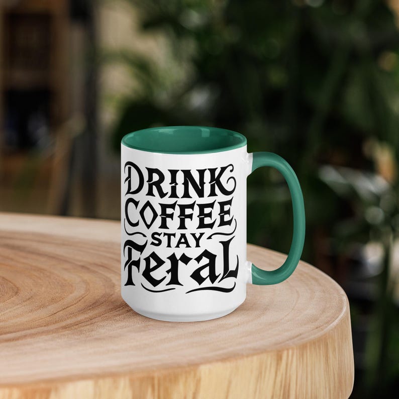 K&ouml;nnte beinhalten: Wei&szlig;er Keramikbecher mit gr&uuml;nem Innenraum und Henkel. Der Becher tr&auml;gt den Text "DRINK COFFEE STAY FERAL" in schwarzer, stilisierter Schrift. Der Becher steht auf einer Holzoberfl&auml;che.