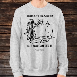 Może przedstawiać: Jasnoszara bluza z czarną grafiką. Grafika przedstawia postać w szacie z laską, gwiazdą i napisem "YOU CAN'T FIX STUPID BUT YOU CAN REZ IT." Poniżej linia na "Player Name."