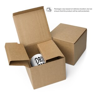 K&ouml;nnte beinhalten: Eine wei&szlig;e Kaffeetasse mit dem Text "DRI" in Schwarz, in einem braunen Karton. Die Box ist ge&ouml;ffnet und zeigt die Tasse und die Schutzverpackung. Ein weiterer geschlossener Karton ist im Hintergrund zu sehen.