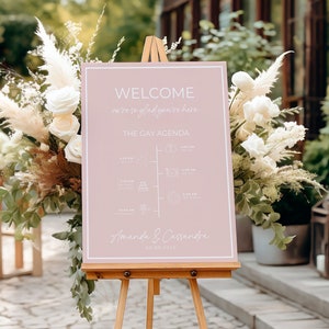 May include: A pink welcome sign with a white easel. The sign says "Welcome, we're so glad you're here. The Gay Agenda." It also includes a timeline of events for the day, and the names "Amanda & Cassandra" at the bottom.