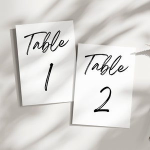 May include: Two white table number cards with black calligraphy. The first card says "Table 1" and the second card says "Table 2".