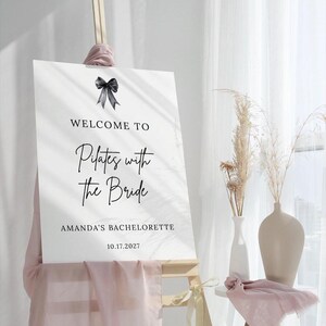 May include: A white sign with black text that reads "WELCOME TO Pilates with the Bride" and "AMANDA'S BACHELORETTE 10.17.2027". A black bow is at the top. The sign is on a wooden easel.