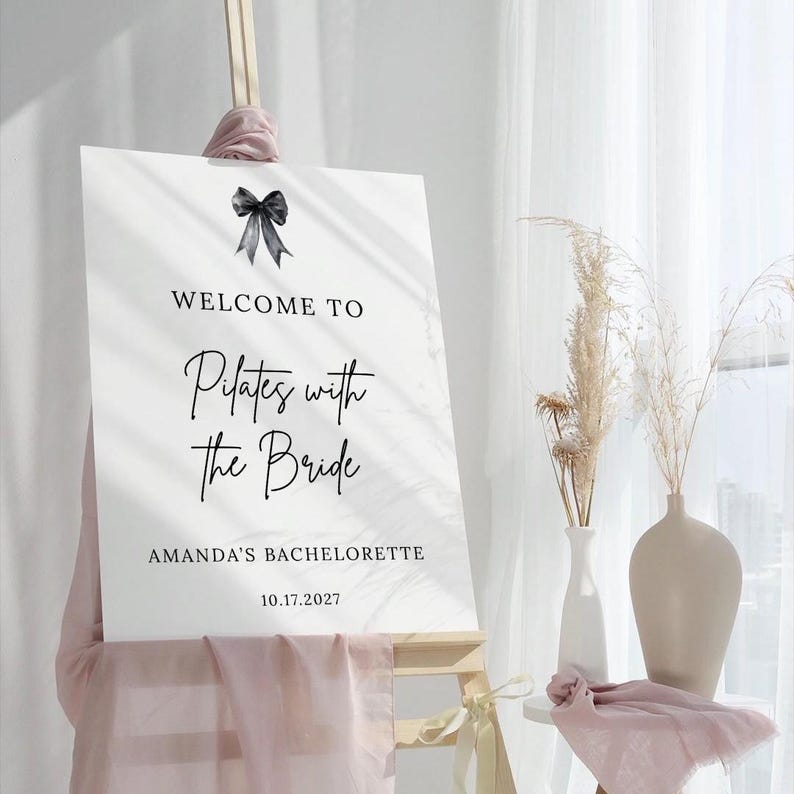 May include: A white sign with black text that reads "WELCOME TO Pilates with the Bride" and "AMANDA'S BACHELORETTE 10.17.2027". A black bow is at the top. The sign is on a wooden easel.