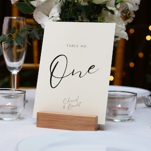 May include: White card table number holder with black calligraphy that says "Table No. One" and "Chanel & Benoit" on a wooden base.