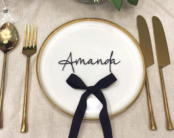 May include: A white ceramic plate with a gold rim featuring the name Amanda in black script lettering and a black ribbon tied in a bow. The plate is flanked by a gold spoon, a gold fork, and two gold knives on a light-colored fabric surface.