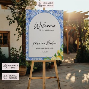 Peut inclure: Panneau de bienvenue de mariage sur un chevalet en bois. Le panneau a un fond blanc avec une bordure à motifs bleus et blancs. Le texte se lit "Bienvenue au mariage de Maria & Pedro" avec la date 21/06/26. Des citrons et de la verdure décorent le bas.