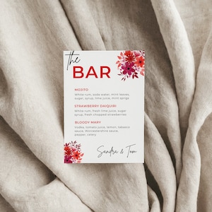 Könnte beinhalten: Ein weißes Schild mit roten floralen Akzenten und dem Text "the BAR" oben. Das Schild listet drei Getränkeauswahlmöglichkeiten auf: Mojito, Erdbeer-Daiquiri und Bloody Mary. Das Schild ist mit "Sandra & Tom" unterschrieben.