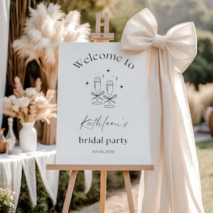 Könnte beinhalten: Ein weißes Willkommens-Schild mit schwarzem Text und einer Grafik von zwei Champagnergläsern mit Schleifen. Der Text lautet "Willkommen zur Brautparty von Kathleen 30.04.2026".