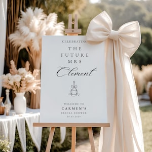 Könnte beinhalten: Ein weißes Schild auf einer Holzstaffelei mit dem Text "Celebrating The Future Mrs Clement" und "Welcome to Carmen's Bridal Shower 30.03.2029". Eine große cremefarbene Schleife ist an der Staffelei befestigt. Dekorative Blumen und Pampasgras sind im Hintergrund.