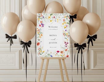 Pode incluir: Uma placa de boas-vindas de casamento em um cavalete de madeira, cercada por balões bege e fitas pretas. A placa apresenta detalhes florais e os nomes "MONA e JUSTIN" com a data "30 de maio de 2018".