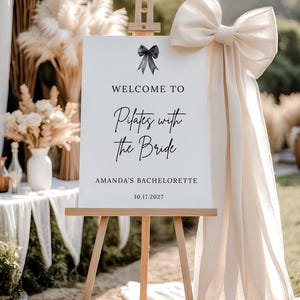May include: A white sign on a wooden easel with the text "WELCOME TO Pilates with the Bride" and "AMANDA'S BACHELORETTE 10.17.2027". A large cream-colored bow and fabric drape to the right. Decorative pampas grass and flowers are in the background.