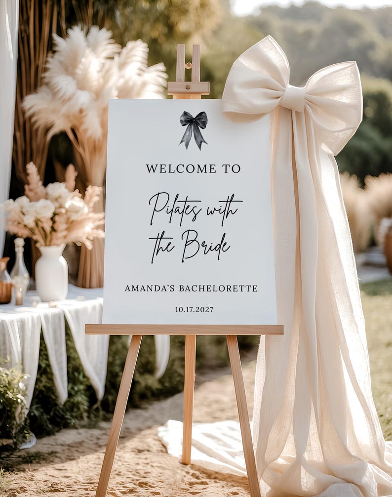 May include: A white sign on a wooden easel with the text "WELCOME TO Pilates with the Bride" and "AMANDA'S BACHELORETTE 10.17.2027". A large cream-colored bow and fabric drape to the right. Decorative pampas grass and flowers are in the background.