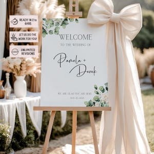 Könnte beinhalten: Eine hölzerne Staffelei zeigt ein Hochzeits-Willkommensschild. Auf dem Schild steht „Willkommen zur Hochzeit von Pamela + Derek“ mit floralen Akzenten. Eine große cremefarbene Schleife ziert die Staffelei. Das Schild enthält auch den Text „Wir freuen uns, dass Sie hier sind“ und das Datum.