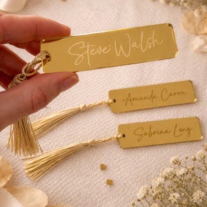 May include: Three rectangular gold-coloured acrylic bookmarks with a reflective finish. Each bookmark features a custom name engraved in a cursive script and is attached to a light beige decorative tassel through a small hole at the top end.