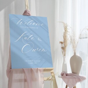 May include: A light blue welcome sign with white calligraphy that reads "Welcome to the wedding of Kate & Owen" and the date "October 23, 2025" and location "Carp, ON".