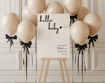 Könnte beinhalten: Ein weißes Schild mit schwarzem Text, auf dem "hello baby" mit einem Herz und "Willkommen zu Melanies Baby Shower 02.27.2027" steht. Das Schild steht auf einem Staffelei und ist von beigefarbenen Ballons mit schwarzen Bändern umgeben.