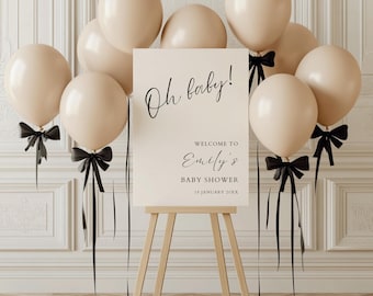May include: A white sign with black text that reads "Oh baby! Welcome to Emily's Baby Shower 18 January 20XX". The sign is propped up on an easel and surrounded by beige balloons with black ribbons.