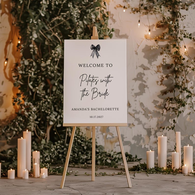 May include: A white sign on a wooden easel with the text "WELCOME TO Pilates with the Bride" and "AMANDA'S BACHELORETTE 10.17.2027". A black bow graphic is at the top. The background includes candles and greenery.