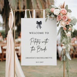 May include: A white sign with the text "WELCOME TO Pilates with the Bride" and "AMANDA'S BACHELORETTE 10.17.2027" is displayed on a gold stand. The sign is decorated with a black bow and floral arrangements.
